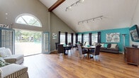 This single-floor space makes a striking architectural statement through its dramatic vaulted ceiling, accented by an exposed dark wood beam that draws the eye upward and amplifies the sense of volume throughout. Warm hardwood flooring runs continuously across the open-concept layout, unifying the kitchen and living areas into one cohesive, flowing environment.

The living area is characterized by its generous proportions and vibrant teal accent wall, which adds bold contrast against the surrounding neutral gray tones. Track lighting mounted along the ceiling ridge provides ample, directed illumination. Multiple windows dressed with dark curtains line the walls, inviting abundant natural light, while a prominent arched window above the French doors frames an outdoor view and further enhances brightness. The French doors themselves open directly to an outdoor area, creating seamless indoor-outdoor connectivity.

The kitchen features dark cabinetry paired with granite-style countertops and a teal backsplash that ties the color palette together. Stainless steel appliances, including a refrigerator, dishwasher, microwave, and range, are neatly integrated. A peninsula counter provides additional workspace and a natural boundary between the kitchen and the open living space, contributing to the efficient, well-organized layout.