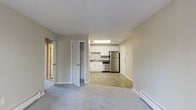 Located at 1445 Bodwell Rd in Manchester, New Hampshire, this single-floor residence offers a clean, practical layout designed for comfortable everyday living. The open living area is finished with soft wall-to-wall carpeting, creating a warm and inviting atmosphere, while neutral-toned walls provide a versatile backdrop throughout the space. The kitchen flows seamlessly from the living area and features light wood-style flooring, crisp white cabinetry, dark countertops, and stainless steel appliances including a refrigerator and range. A stainless steel sink and dishwasher complete the kitchen's functional setup, all illuminated by overhead fluorescent lighting. A closet with bi-fold doors provides generous built-in storage directly off the main living space, keeping the layout organized and efficient. A hallway connects to additional rooms, offering a sense of separation between living and private areas. Baseboard heating is present throughout, ensuring reliable warmth during New Hampshire's colder seasons. The overall aesthetic is clean and understated, making the space easy to personalize and move into.