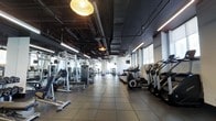 This modern fitness facility offers a thoughtfully designed workout environment that balances industrial aesthetics with functional practicality. The space features an exposed ceiling with visible ductwork and piping, painted in a dark tone that contrasts sharply with the bright, linear LED lighting strips running the length of the room. Complementing the contemporary lighting scheme is a distinctive pendant light fixture that adds a unique design element overhead.

The flooring consists of large-format tiles, providing a clean and durable surface throughout the main workout area. Natural light filters in through tall windows along one side of the space, contributing to an open and energizing atmosphere.

The facility is organized into clearly defined zones. The strength training area is stocked with a variety of cable machines, free weights, and resistance equipment arranged for ease of access and movement. On the opposite side, a dedicated cardio zone houses a row of elliptical trainers and treadmills, positioned to allow users to face the windows. Mirrored walls throughout the space enhance the sense of openness and allow members to monitor their form during workouts.

Overall, the gym presents a spacious, well-lit, and comprehensively equipped environment suitable for a wide range of fitness goals and training styles.