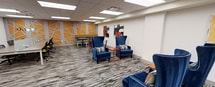 This single-floor space is a thoughtfully designed lounge or common area that blends bold aesthetics with functional comfort. The room is immediately eye-catching, anchored by a striking accent wall featuring a geometric orange-and-white pattern that adds energy and visual depth to the space. Complementing this are contemporary abstract art pieces displayed on adjacent walls, lending the area a creative and inspiring atmosphere.

The flooring consists of patterned carpet in neutral gray tones with subtle striping, providing a grounded contrast to the vibrant décor above. Seating is a standout feature, with plush royal blue wingback chairs accented by colorful striped throw pillows arranged around small orange side tables, creating intimate conversation areas throughout the room. Additional seating options include modern task chairs and a casual café-style table and chair grouping tucked near the accent wall, offering versatile use for both relaxed socializing and informal work.

Overhead, a suspended ceiling with recessed panel lighting ensures the space is evenly and brightly illuminated throughout. The overall design palette — combining deep blues, bold oranges, and crisp whites — creates a welcoming, dynamic environment well-suited for collaboration, relaxation, or informal gatherings.
