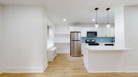 This thoughtfully renovated single-floor unit showcases a clean, modern aesthetic defined by crisp white walls, light hardwood flooring, and recessed lighting throughout. The open-concept layout connects the living area and kitchen seamlessly, creating a bright and airy atmosphere. Multiple windows fitted with blinds allow natural light to fill the space, complementing the warm tones of the flooring.

The kitchen is a standout feature, equipped with white shaker cabinetry accented by matte black hardware, quartz countertops, and a distinctive blue herringbone-pattern tile backsplash. Stainless steel appliances — including a gas range, microwave, refrigerator, and dishwasher — are neatly integrated alongside a center island with pendant lighting overhead. A built-in shelving nook adjacent to the kitchen offers additional functional space.

The living area is generously proportioned, with radiant baseboard heating units positioned beneath the windows. Bedrooms are calm and understated, with ample natural light and continued hardwood flooring. The bathroom features geometric patterned wall tile surrounding a soaking tub, slate-toned floor tiles, a natural light window, and matte black fixtures, adding a refined, contemporary touch.