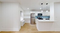 This thoughtfully renovated single-floor unit showcases a clean, modern aesthetic defined by crisp white walls, light hardwood flooring, and recessed lighting throughout. The open-concept layout connects the living area and kitchen seamlessly, creating a bright and airy atmosphere. Multiple windows fitted with blinds allow natural light to fill the space, complementing the warm tones of the flooring.

The kitchen is a standout feature, equipped with white shaker cabinetry accented by matte black hardware, quartz countertops, and a distinctive blue herringbone-pattern tile backsplash. Stainless steel appliances — including a gas range, microwave, refrigerator, and dishwasher — are neatly integrated alongside a center island with pendant lighting overhead. A built-in shelving nook adjacent to the kitchen offers additional functional space.

The living area is generously proportioned, with radiant baseboard heating units positioned beneath the windows. Bedrooms are calm and understated, with ample natural light and continued hardwood flooring. The bathroom features geometric patterned wall tile surrounding a soaking tub, slate-toned floor tiles, a natural light window, and matte black fixtures, adding a refined, contemporary touch.