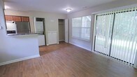 This single-floor residence offers a clean, functional layout with a warm and welcoming ambiance throughout. The main living area is finished with smooth hardwood-style flooring that extends seamlessly across the open space, lending a cohesive and polished look. Neutral-toned walls keep the interior bright and versatile, complementing a variety of décor styles.

Natural light fills the living area through a large window fitted with horizontal blinds and a sliding glass door dressed with vertical blinds, which opens to a view of lush, mature greenery outdoors. This connection to the outside enhances the overall sense of openness within the space.

A built-in closet with double doors provides convenient storage directly within the living area, while a compact white storage cabinet nearby adds additional utility. The in-unit stacked washer and dryer, neatly tucked into a dedicated nook adjacent to the kitchen area, offer practical everyday convenience without sacrificing space. A partial view of the kitchen reveals stone-look countertops, suggesting a functional and well-appointed cooking area. Overhead, a flush-mount ceiling light provides clean, even illumination throughout. This residence balances simplicity and practicality in a tidy, move-in-ready package.
