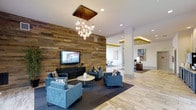 This thoughtfully designed single-floor space makes a strong first impression with its blend of modern aesthetics and warm, inviting materials. The centerpiece of the lounge area is a dramatic floor-to-ceiling reclaimed wood accent wall, which anchors the room and provides rich texture against the clean white walls and structural columns. Mounted above a sleek black media console, a flat-screen television serves as a functional focal point for the seating arrangement below.

The lounge is furnished with a curated collection of contemporary blue upholstered armchairs and a low-profile sofa, arranged around a minimalist round coffee table atop a deep charcoal area rug. Overhead, a statement chandelier featuring clustered globe bulbs suspended from copper rods adds a sculptural, artistic element to the space.

Light wood-toned flooring extends throughout, contributing to an open, airy feel. Recessed lighting throughout the ceiling complements the natural tones of the materials. Beyond the lounge, an adjacent open area is visible, featuring additional seating, accent artwork on the walls, and warm accent lighting — suggesting a versatile, multi-purpose layout ideal for gathering, collaboration, or relaxation.