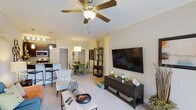 Located at 1000 Antietam Creek Dr in Fayetteville, NC, this single-floor residence offers a thoughtfully designed open-concept layout that connects the living, dining, and kitchen areas in a cohesive and functional arrangement. The living area is appointed with plush carpeting, crown molding, and a ceiling fan with integrated lighting that enhances both comfort and style. A large flat-panel television mount anchors one wall, complemented by generous space for furniture and décor. The dining area sits adjacent to the living room, illuminated by a modern pendant light fixture that adds a warm, inviting ambiance. The kitchen features dark cabinetry, stainless steel appliances including a built-in microwave, and a breakfast bar with seating — ideal for casual dining or entertaining. The open sightlines throughout the main living space create a sense of spaciousness and connectivity. Neutral wall tones and consistent flooring choices contribute to a clean, modern aesthetic throughout. The overall design balances practicality with comfort, making this a well-appointed residence suited for everyday living in Fayetteville, NC.