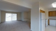 This single-floor space offers a functional and understated layout designed for comfortable everyday living. The main living area is open and airy, featuring neutral gray carpeting that extends throughout and smooth white walls that lend a clean, minimalist aesthetic. A sliding glass door along one wall fills the space with natural light while providing direct access to the outdoors, offering views of surrounding greenery.

A partial wall with a railing separates the living area from a lower-level staircase, adding architectural depth and a sense of layered space. The transition between areas is defined by a subtle structural beam overhead, contributing to the overall layout without interrupting the open feel.

Adjacent to the living area, the kitchen is partially visible and includes warm-toned wood cabinetry positioned above, paired with a full-size white refrigerator. The cabinetry provides ample storage, while the neutral color palette maintains visual continuity with the rest of the space.

Overall, the space presents a straightforward, well-maintained interior that balances practicality with a light, open atmosphere suited to a variety of living arrangements.