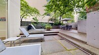 Located at 1255 Pendrell St in Vancouver's West End, this ground-level outdoor amenity space offers a generous and well-appointed patio environment framed by mature trees and established greenery. The area is finished with aggregate concrete paving laid out across multiple levels connected by low steps, creating distinct zones for relaxation and outdoor dining.

A dedicated lounging area features modern sun chairs arranged to take advantage of the open sky, while a separate dining zone includes a patio table with chairs and a large umbrella for shade. A gas BBQ station is conveniently positioned nearby, making the space well-suited for outdoor entertaining. Lush planters and raised garden beds line the perimeter, adding natural softness to the concrete hardscape.

Architecturally, the space is enclosed by smooth concrete walls and incorporates distinctive glass block panels that allow diffused light to pass between the outdoor and indoor areas. Large sliding glass doors connect the patio directly to an interior space that appears to include an indoor pool, enhancing the overall recreational offering.

The mature tree canopy overhead provides a sense of privacy and seclusion, creating a tranquil urban retreat. The combination of thoughtful zoning, greenery, and modern amenities makes this outdoor space a functional and inviting extension of the building's residential facilities.