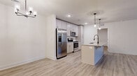 This well-appointed single-story home offers three bedrooms, two bathrooms, and a thoughtfully designed layout that maximizes both functionality and comfort. The open-concept kitchen and living area serves as the heart of the home, featuring light wood-style flooring throughout, recessed lighting, and a stylish chandelier. The kitchen is equipped with stainless steel appliances — including a French door refrigerator, range, and microwave — complemented by white shaker-style cabinetry, granite countertops, and a central island with pendant lighting and a built-in sink, providing ample prep and casual dining space. The living room is bright and airy, benefiting from natural light through multiple windows, a ceiling fan, and direct access to an exterior door. The primary bedroom is generously sized and includes a walk-in closet outfitted with wire shelving and basket storage. The primary bathroom features a granite-topped vanity with white cabinetry, a glass-enclosed walk-in shower, and wood-style tile flooring. Additional bedrooms are carpeted and versatile in use. The home also includes a utility room and a second closet for added storage. The clean, neutral color palette and modern fixtures throughout create a bright, cohesive living environment.