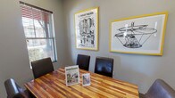 Study Room 2 is a focused, private meeting space designed to facilitate productive sessions in a comfortable and visually engaging environment. The room centers around a rich, warm-toned butcher block-style wooden table surrounded by dark leather chairs, providing seating for a small group. The flooring is a neutral carpet that complements the room's gray-painted walls, lending a calm and professional atmosphere throughout.

A standout feature of the space is the pair of bold, yellow-framed art prints displayed prominently on one wall — one depicting an intricate illustrated scene titled "The Thought Machine," and the other showcasing a detailed sketch of a flying apparatus, both adding a creative and intellectually stimulating character to the room. A wall-mounted flat-screen television is positioned on the adjacent wall, making the space suitable for presentations or collaborative viewing.

Natural light filters in through a window fitted with horizontal blinds, offering a view of the surrounding exterior while allowing light control as needed. A glass-paneled door provides easy access and a sense of openness. The room is compact yet efficiently arranged, making it well-suited for small group study sessions, tutoring, or private meetings.