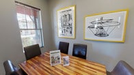 Study Room 2 is a focused, private meeting space designed to facilitate productive sessions in a comfortable and visually engaging environment. The room centers around a rich, warm-toned butcher block-style wooden table surrounded by dark leather chairs, providing seating for a small group. The flooring is a neutral carpet that complements the room's gray-painted walls, lending a calm and professional atmosphere throughout.

A standout feature of the space is the pair of bold, yellow-framed art prints displayed prominently on one wall — one depicting an intricate illustrated scene titled "The Thought Machine," and the other showcasing a detailed sketch of a flying apparatus, both adding a creative and intellectually stimulating character to the room. A wall-mounted flat-screen television is positioned on the adjacent wall, making the space suitable for presentations or collaborative viewing.

Natural light filters in through a window fitted with horizontal blinds, offering a view of the surrounding exterior while allowing light control as needed. A glass-paneled door provides easy access and a sense of openness. The room is compact yet efficiently arranged, making it well-suited for small group study sessions, tutoring, or private meetings.