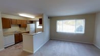 This single-floor unit offers an open and functional layout that connects the kitchen and living area in a cohesive, easy-to-navigate design. The living area is finished with wall-to-wall carpeting in a neutral tone, contributing to a warm and comfortable atmosphere. A large window dominates one wall, allowing abundant natural light to fill the space while framing a view of surrounding greenery outdoors.

The kitchen is partially separated from the living area by a half-wall breakfast bar, creating a natural divide while maintaining an open feel between the two spaces. The kitchen features warm wood-toned cabinetry, a white refrigerator, and tile flooring that contrasts with the carpeted living area. An overhead fluorescent light fixture provides practical task lighting above the kitchen workspace, and a secondary window within the kitchen adds additional natural light to the cooking area.

The neutral wall tones throughout the unit create a clean, versatile backdrop suitable for a variety of interior styles. The overall ambiance is bright and straightforward, with a practical, well-organized layout ideal for everyday living.
