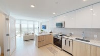 Located along Hollywood Boulevard in Hollywood, FL, this contemporary residence offers a thoughtfully designed single-floor layout with a bright, modern aesthetic. The open-concept kitchen and living area serve as the heart of the space, where high-gloss white upper cabinetry pairs with warm natural wood lower cabinets and a striking veined quartz waterfall island countertop. A neutral tile backsplash and full suite of stainless steel appliances — including a range, microwave, and refrigerator — complete the kitchen. The living area flows seamlessly from the kitchen, bathed in abundant natural light from floor-to-ceiling sliding glass doors that open to a balcony with expansive views of the Hollywood cityscape. Light-toned porcelain tile flooring runs throughout the common areas, enhancing the airy ambiance. A compact dining area with a round marble-top table adds a refined touch alongside the living space. The bedroom is a calm retreat, featuring recessed lighting, a window with outdoor views, and a neutral color palette that promotes rest. The bathroom is clean and functional, finished with light wood vanity cabinetry and a full tub-shower combination. Recessed lighting throughout and crisp white walls unify the space with a polished, contemporary feel.