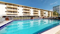 This multi-story building presents a classic Florida-style architectural design, characterized by its cream-colored exterior, decorative lattice screens, and private balconies extending from each unit across multiple floors. The building's facade reflects a mid-century tropical aesthetic, with repeating geometric detailing that adds visual interest while providing shade and privacy to residents.

A standout feature of this property is the expansive outdoor swimming pool situated at the base of the building. The generously sized pool showcases vivid turquoise water and is enclosed by a wrought-iron fence, providing both safety and an open sightline to the building. Potted flowering plants accent the pool deck, adding color and a welcoming atmosphere to the outdoor space.

The ground-floor level includes covered common areas with dark awnings, suggesting shared amenity or lobby spaces accessible to residents. The surrounding environment reflects a warm, sunny climate, enhanced by the presence of palm trees and clear blue skies, contributing to the overall resort-like ambiance of the property.