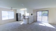 This single-floor residence features an open-concept design that connects the kitchen and living area into one cohesive, light-filled space. The kitchen is equipped with white cabinetry, a stainless steel refrigerator, and a built-in range, complemented by a peninsula that doubles as a breakfast bar and incorporates open shelving on the living room side — a functional detail that adds storage while maintaining visual openness between the two spaces. The living area is finished with neutral wall-to-wall carpeting that contributes to a warm, comfortable atmosphere. Large sliding glass doors lead to an outdoor area, allowing generous natural light to fill the interior throughout the day. Additional windows positioned near the kitchen further enhance the bright, airy feel of the space. A ceiling fan is present in the living area, supporting year-round comfort. The overall color palette — whites, beiges, and soft neutrals — lends the space a clean, understated character. The layout is practical and well-organized, making efficient use of the available floor plan on a single level.