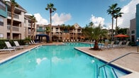 This amenity space centers around a generously sized outdoor swimming pool with crystal-clear turquoise water, framed by a wide pool deck lined with lounge chairs and shaded by tall, tropical palm trees. A covered canopy area provides additional shade for residents, while the surrounding multi-story buildings create an enclosed, resort-like atmosphere. The deck is finished with tasteful hardscape tiling and features landscaped planting beds that add a touch of greenery to the outdoor environment.

Access to the interior facility is provided through a glass-paneled door that offers clear sightlines to the pool area and courtyard beyond, with a secure iron railing fence delineating the pool zone. Inside, the clubhouse-style interior features light wood-look flooring, tray ceilings with recessed lighting, and an open layout that flows from the entry toward a central island with bar seating and a well-equipped kitchen area visible in the background. Framed artwork adorns the neutral-toned walls, lending a polished, welcoming aesthetic to the space. Clearly marked, ADA-compliant restroom facilities with dedicated men's and women's entrances are conveniently located within the interior. The combination of indoor comfort and expansive outdoor amenities makes this a comprehensive and functional community gathering space.