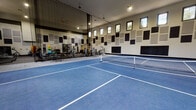 This single-floor athletic facility is designed to accommodate a wide range of fitness and sports activities within one expansive, open interior. The centerpiece of the space is a full-size indoor court with a vibrant blue interlocking tile surface, marked for both pickleball and tennis, complete with portable nets. Soaring ceilings provide the vertical clearance needed for active play, while a row of clerestory windows along the upper walls allows natural light to filter in throughout the day. A retractable curtain divider separates the court area from the fitness zone, offering flexibility in how the space is used. The fitness area is thoroughly equipped with a wide selection of cardio machines — including treadmills, ellipticals, and stationary bikes — all positioned in front of wall-mounted monitors. A dedicated strength training section features cable machines, weight benches, and a multi-functional rack stocked with medicine balls, resistance bands, and accessories. Throughout the facility, acoustic foam panels are strategically arranged in a grid pattern across the white block walls, contributing to sound control and lending the space a modern, purposeful aesthetic. Rubber flooring in the fitness zone ensures durability and safety during training sessions.