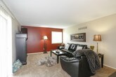 This single-story home offers a well-organized layout across several distinct living spaces. The living room serves as a central gathering area, furnished with ample seating and carpeted flooring, with a ceiling ventilation unit overhead and neutral walls that create a versatile backdrop. Adjacent to it, the dining area features a ceiling fan with lighting, a window dressed with sheer red curtains that allow natural light, and direct proximity to the kitchen for convenient entertaining.

The kitchen is equipped with white cabinetry, tile flooring, and black appliances including a refrigerator, dishwasher, and microwave, providing a functional and clean workspace.

The primary bedroom is a spacious retreat with an accent wall, carpeted flooring, and a window with decorative draping. It connects directly to an en-suite vanity area featuring a white cabinetry base, a wide countertop with an undermount sink, and a large mirror, which leads further into a full bathroom. Additional bedrooms offer carpeted floors, natural light from windows, and sufficient space for a bed and desk setup.

A carpeted hallway connects the secondary bedrooms to a shared full bathroom, complete with a tub-shower combination and tile flooring, completing this functional and cohesive home.