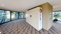 The Club View Parcel Locker room is a thoughtfully designed, single-floor space that serves as a dedicated package management area for residents. The room features a distinctive curved architectural layout, with sweeping walls that guide the eye naturally through the space. Large floor-to-ceiling windows line the exterior walls, flooding the interior with abundant natural light and offering pleasant views of the surrounding landscaping and mature trees outside.

The flooring throughout consists of a patterned carpet in neutral tones with a subtle circular motif, complemented by dark wood baseboards that provide a clean contrast against the warm beige walls. Recessed ceiling lighting supplements the natural light, ensuring the space remains bright and functional at all times. The textured popcorn ceiling adds a classic finish to the room.

The centerpiece of the space is a modern Parcel Pending smart locker system, branded with Club View's logo. This large, white locker unit offers multiple compartments of varying sizes, equipped with an electronic interface for secure, convenient package retrieval. An emergency exit sign is clearly visible, reflecting attention to safety. The overall ambiance is clean, welcoming, and practical, making package pickup a straightforward and comfortable experience for residents.