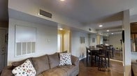 This single-floor space presents an open-concept layout that connects the living and dining areas in a functional and cohesive arrangement. The living area is anchored by a comfortable upholstered sofa accented with decorative pillows, set against neutral gray walls that lend a calm, understated tone to the space. Overhead, recessed lighting and a ceiling-mounted HVAC vent ensure year-round comfort, while a wall-mounted HVAC return adds to the practical infrastructure of the home. Rich, dark hardwood flooring runs throughout, providing a warm contrast to the lighter wall tones and tying the spaces together visually. Decorative wall art featuring feather motifs adds a subtle, stylistic touch to the living area. Transitioning seamlessly into the dining zone, a dark wood dining table is surrounded by matching chairs, creating a defined yet open eating area. A large mirror on the adjacent wall enhances the sense of space by reflecting light and depth. A doorway visible from the living area suggests additional rooms beyond, contributing to the overall functional flow of this well-appointed, single-level residence.