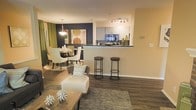 This well-appointed single-floor residence showcases an open-concept layout that effortlessly connects the living, dining, and kitchen areas into one cohesive and functional space. The living area features rich, dark hardwood-style flooring and is anchored by a wood coffee table and a plush accent chair, creating a comfortable and inviting atmosphere. A textured area rug adds warmth and definition to the seating zone.

The kitchen is positioned at a half-wall breakfast bar, complete with dark countertops and two industrial-style bar stools that provide casual seating and a sense of separation without closing off the space. White cabinetry lines the kitchen walls, complemented by a mosaic tile backsplash and track lighting overhead that illuminates the workspace efficiently.

The dining area flows naturally from the kitchen and living space, highlighted by a bold navy accent wall that serves as a striking focal point. A pendant light fixture hangs above the dining table, casting a warm glow over the seating arrangement of elegantly styled chairs. Olive green curtains frame a window, adding a rich pop of color and connecting the interior palette cohesively. Together, these spaces form a bright, modern, and thoughtfully designed interior ideal for everyday living and entertaining.