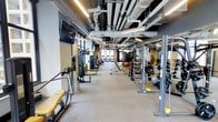 This fitness facility offers a comprehensive workout environment designed to accommodate a variety of training needs. The space features an open, elongated floor plan with a neutral-toned carpet flooring that provides comfort underfoot throughout the workout area. The ceiling is deliberately left exposed, showcasing industrial-style ductwork, piping, and linear LED lighting that runs the length of the space, contributing to a modern, utilitarian aesthetic while ensuring consistent illumination across the entire facility.

Strength training equipment lines both sides of the room, including cable machines, weight racks stocked with dumbbells and barbells, adjustable benches, and resistance machines. Stability balls and additional functional training accessories are also visible, supporting a well-rounded training program. A wall-mounted flat-screen television is positioned for easy viewing during workouts. Toward the far end of the space, natural light filters in through large windows, adding brightness and an open feel to the facility. The overall layout is organized and spacious, allowing for efficient movement between equipment stations and accommodating multiple users simultaneously.