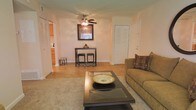 This single-floor residence offers a welcoming and functional layout designed for comfortable everyday living. The living room serves as the heart of the space, featuring a blend of hardwood flooring and plush carpet that adds warmth and texture underfoot. Neutral, warm-toned walls create a calm and inviting ambiance throughout.

A ceiling fan with integrated lighting is centrally positioned, ensuring year-round comfort and consistent airflow. The living area is well-proportioned, accommodating a sofa and a dark-finished coffee table with glass inlay, while a compact bar-height table with stools provides a casual dining or entertaining option beneath a framed wall piece.

Ample storage is evident through multiple closet doors visible throughout the space, offering practical organization solutions. The open sightline into the kitchen reveals wood-toned cabinetry and standard appliances, suggesting a seamlessly connected layout that supports ease of movement between rooms.

The overall ambiance is warm and understated, with a traditional aesthetic carried through consistent furnishings and finishes. The space is well-suited for those seeking a practical and comfortable single-floor living environment with efficient use of space throughout.