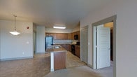 This single-floor space centers around an open-concept kitchen that flows naturally into the adjacent living area. The kitchen is well-appointed with warm medium-toned wood cabinetry lining the walls, offering generous storage throughout. A peninsula-style island with a granite-look countertop and an integrated sink serves as a functional focal point, providing additional workspace and a natural divider between the kitchen and the carpeted living space. Black appliances, including a refrigerator, range, and built-in microwave, offer a clean contrast against the lighter countertops and cabinetry. The flooring in the kitchen area consists of tile in a neutral tone, transitioning to carpet in the adjoining living space. Recessed and flush-mounted ceiling lighting provides consistent illumination throughout the kitchen. Two paneled doors are visible from the kitchen—one leading to what appears to be a closet or pantry, and another opening to an additional room, suggesting functional separation of spaces. The overall design is practical and cohesive, with a neutral color palette of beige, gray, and warm wood tones creating a welcoming, understated ambiance throughout.
