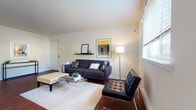 This single-floor residence offers a thoughtfully designed layout that balances comfort and functionality across its key living spaces. The living room serves as the heart of the home, showcasing rich hardwood flooring, neutral walls, and ample natural light streaming through slatted blinds. The space accommodates a modern seating arrangement, a glass-top coffee table, and a decorative wall shelf, creating an inviting and contemporary atmosphere. Adjacent to the living room, a dedicated dining area features the same warm hardwood floors, a round glass-top dining table with upholstered chairs, and a pendant light overhead, with a window providing natural light. The galley-style kitchen is efficiently organized with dark cabinetry lining both sides, light countertops, white appliances including a range and dishwasher, and tile flooring. The bedroom is carpeted and well-proportioned, offering a comfortable retreat with natural light from a window. Two bathrooms serve the residence, each finished with tile flooring and warm wood vanity cabinetry topped with light counters and illuminated mirrors. Together, the spaces present a cohesive, clean aesthetic with consistent warm-toned finishes throughout.