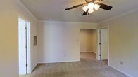This single-floor residence presents a clean, functional layout designed for comfortable everyday living. The main living area is finished with wall-to-wall carpeting in a neutral beige tone, lending a warm and cohesive feel throughout the space. Crown molding runs along the ceiling perimeter, adding a subtle architectural detail that elevates the room's character. A ceiling fan with an integrated light fixture is centrally positioned, providing both air circulation and ambient lighting. The walls are painted in a soft, neutral tone that enhances the sense of openness and pairs well with the light-colored carpeting. A wall-mounted ventilation grille is visible, indicating a climate control system built into the home. An open passageway connects the main living area to an adjoining room, allowing for a seamless flow between spaces. A white paneled door leads to an additional room or closet, contributing to the overall practicality of the floor plan. Electrical outlets are conveniently placed throughout the walls. The space is clean, well-maintained, and move-in ready, offering a straightforward and adaptable canvas well-suited for a variety of furnishing arrangements and personal styles.