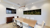 This single-floor professional space is designed with a clean, contemporary aesthetic that balances functionality with thoughtful design details. The centerpiece of the space is a well-appointed conference room, anchored by a large rectangular wood-finished meeting table surrounded by sleek white ergonomic chairs. A wall-mounted flat-screen display and a rich dark wood credenza provide practical AV and storage solutions, while a curved wood-accented pendant light fixture adds warmth overhead.

A standout architectural feature is a large recessed living moss wall installation, showcasing lush preserved greenery and sculptural driftwood branches, bringing a striking natural element into the otherwise crisp, white-walled interior. Recessed and ambient lighting throughout ensures a bright, professional atmosphere.

Floor-to-ceiling glass partitions along one side of the conference room provide visual connectivity to adjacent areas, including a modern kitchen and lounge space with pendant lighting, a central island, and bar-height seating. Tall windows allow natural light to flow in from outside, where an outdoor terrace with pergola elements is visible, further enhancing the openness of the space. Interior plantings of snake plants add additional greenery throughout, contributing to a clean, biophilic design sensibility across the entire floor.