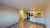 This single-floor unit offers a thoughtfully designed layout that integrates the kitchen and living areas into one cohesive open-concept space. The kitchen is a standout feature, equipped with stainless steel appliances — including a refrigerator, electric range, and dishwasher — paired with crisp white cabinetry, granite countertops, and a decorative geometric tile backsplash. A peninsula counter provides additional workspace and casual seating. Track lighting overhead ensures the kitchen is well-illuminated. Wood-look flooring runs throughout the main living area, lending a clean, contemporary feel.

The living area is open and versatile, with neutral wall tones and a hallway leading to the private spaces of the unit. The bedroom features plush carpeting, a ceiling fan with light fixture, a window with blinds that allows natural light and outdoor views, and a baseboard heating unit. A wall-mounted HVAC unit provides additional climate control in the living space.

The bathroom is finished with a full soaking tub and shower combination, accented by white subway tiles and a vertical decorative tile strip in a warm taupe picket pattern, complemented by brushed bronze fixtures for an elevated touch.