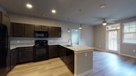 This single-story residence in Kyle, Texas, presents a cohesive, modern interior defined by light wood-style flooring and neutral gray walls throughout. The open-concept kitchen serves as the functional centerpiece of the home, featuring rich dark cabinetry that contrasts sharply with light granite countertops. A peninsula with an undermount sink and pendant lighting above extends into the living area, creating a natural gathering point. Black appliances, including a refrigerator, range, and built-in microwave, complete the kitchen's bold, contemporary aesthetic. Recessed lighting provides consistent illumination across the space.

The layout connects the kitchen seamlessly to the living and hallway areas, which offer additional closet storage and clear sightlines throughout. Multiple bedrooms share a consistent design language: light flooring, ceiling fans, and windows fitted with blinds that allow natural light to filter in throughout the day.

The bathrooms are well-appointed, with one featuring a granite-topped vanity with dark cabinetry and a mirror, and another offering a full tub-shower combination with built-in shelving. Both bathrooms continue the light flooring found throughout the home, maintaining a unified and clean aesthetic across the entire residence.