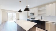 This thoughtfully designed one-bedroom, one-bathroom unit delivers a clean, contemporary aesthetic across a single floor. The kitchen serves as the centerpiece of the space, featuring a striking two-tone design with crisp white upper cabinetry and dark lower cabinets, a geometric tile backsplash, light quartz countertops, and a central island topped with matching quartz. A full suite of stainless steel appliances — including a French door refrigerator, electric range, microwave, and dishwasher — is complemented by two bold black dome pendant lights suspended from brass fixtures, adding a modern industrial touch. Light hardwood flooring runs throughout the main living areas, enhancing the bright, airy atmosphere. The living area benefits from a sliding glass door that opens to a private balcony, filling the space with natural light and offering outdoor views. The bedroom is carpeted for comfort, features a large window with ample natural light, and includes access to a dedicated closet. The bathroom is finished with dark wood cabinetry, a white countertop vanity, matte black hardware, and a full soaking tub with shower. Neutral warm-toned walls and consistent modern fixtures tie the entire unit together cohesively.
