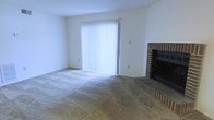 This single-floor space presents a straightforward and functional layout centered around a comfortable living area. The room is finished with wall-to-wall carpet in a neutral tan tone, providing a warm and practical foundation throughout. A standout feature of the living area is the classic brick fireplace positioned in the corner, complete with a raised brick hearth that adds a traditional architectural element and serves as a natural focal point. The fireplace surround is constructed in a rich, dark brick, lending a sense of character and warmth to the otherwise neutral space. A large sliding glass door dominates the adjacent wall, fitted with vertical blinds that allow for adjustable natural light and offer access to the outdoors. The walls are finished in a clean, bright white, contributing to an airy and open feel. The overall ambiance is simple and unfussy, making it an adaptable space well-suited to a variety of furniture arrangements and personal styles. The single-floor layout ensures ease of movement throughout, and the combination of the fireplace and sliding door provides both comfort and connectivity to the exterior environment.