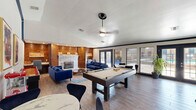 Located at 3011 East Gore Boulevard in Lawton, Oklahoma, this single-floor space combines a fully equipped kitchen with a versatile open living area, offering a comfortable and visually striking environment. The kitchen is a standout feature, showcasing bold navy blue cabinetry adorned with gold hardware, white marble-look countertops, a double stainless steel sink, and full-size appliances including a refrigerator and dishwasher. A large kitchen island with a rich wood base and seating for multiple guests serves as the centerpiece, illuminated by warm Edison-style pendant lights that enhance the gold-and-blue color scheme. The open living area flows naturally from the kitchen and is anchored by a classic brick fireplace flanked by warm wood-paneled walls, adding rustic character to the contemporary design. Vaulted ceilings in the living space create an airy, expansive feel. The room accommodates plush navy blue sofas, a shuffleboard table, a pool table, and a coffee bar station, making it well-suited for social gatherings. A built-in desk nook provides a dedicated workspace. Wood-look tile flooring runs throughout, tying the spaces together cohesively. French doors invite natural light and offer views of the surrounding streetscape, further enhancing the open, welcoming atmosphere of this multi-functional space.