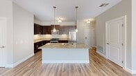 This well-appointed one-bedroom, one-bathroom unit in Hamden, CT offers a thoughtfully designed single-floor layout that maximizes functionality and comfort. The open-concept kitchen and living area serves as the heart of the home, showcasing rich dark-stained cabinetry, granite countertops, and a full suite of stainless steel appliances including a gas range and refrigerator. A central island with pendant lighting adds both workspace and visual appeal, making the kitchen equally suited for cooking and entertaining. Warm hardwood-style flooring flows seamlessly throughout the entire unit, complementing the neutral gray walls and bright white trim that give the space a clean, modern aesthetic. The living area benefits from generous windows dressed with horizontal blinds, allowing ample natural light to fill the room. The spacious bedroom features a large walk-in closet and windows on multiple walls, further enhancing the bright and airy feel. The full bathroom includes a tub-shower combination with accessibility grab bars and a window for natural ventilation. A connecting hallway ties the rooms together efficiently. The overall ambiance is fresh, neutral, and move-in ready, ideal for those seeking a comfortable and contemporary residence in Hamden.