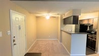 This single-floor space features an open-concept layout that seamlessly connects the living area to the kitchen. The living room is finished with wall-to-wall carpeting in a neutral tone, complemented by beige walls and clean white baseboards that contribute to a cohesive, understated aesthetic. A ceiling fan with an integrated light fixture is centrally positioned overhead, providing both airflow and ambient lighting. The textured popcorn ceiling adds a classic touch to the space.

Transitioning toward the kitchen, a raised breakfast bar with a dark countertop creates a natural division between the two areas while maintaining the open feel. Dark-toned upper cabinetry lines the kitchen walls, offering ample storage, and a sink is visible along the counter. The entryway, marked by a white paneled door, is positioned to the left and features dark flooring that contrasts with the carpeted living area. HVAC vents are integrated into the ceiling, ensuring climate control throughout. Overall, the space presents a functional and straightforward layout suited to everyday living.