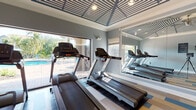Located at 10200 Falcon Pine Blvd in Orlando, Florida, this fitness center is a well-appointed, single-floor facility designed to support a complete cardio and exercise experience. The space is immediately distinguished by its striking architectural ceiling, featuring a geometric pattern of blue and white striped panels accented by crown molding and recessed lighting, lending the room a refined yet energetic character.

The flooring is a standout design element, composed of multicolored vinyl planks in shades of blue, tan, and wood-tone arranged in a bold patchwork pattern that adds vibrancy to the space. Large windows and glass doors line the walls, flooding the interior with natural light and offering direct views of an outdoor pool area surrounded by lush Florida landscaping and palm trees — creating an inviting, resort-like atmosphere.

The facility is equipped with multiple treadmills and elliptical machines from Matrix, catering to a range of cardio workout preferences. Full-length mirrors along the walls visually expand the space and allow users to monitor their form. A ceiling fan contributes to air circulation, enhancing comfort during workouts. An adjacent room, visible through glass-paneled doors, suggests additional amenity space nearby. Overall, this fitness center combines functional equipment with thoughtful design to deliver an elevated exercise environment.