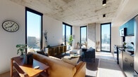 This single-floor unit showcases an industrial-modern aesthetic defined by its signature exposed concrete ceiling, which spans the open-concept living and kitchen area and adds raw, textural character throughout. Floor-to-ceiling black-framed windows wrap the living space, flooding the interior with abundant natural light and framing panoramic city skyline views from an elevated vantage point. Light wood flooring anchors the space, providing a warm contrast to the concrete overhead.

The living area is generously proportioned, accommodating a leather sofa, velvet accent chairs, a gold-accented media console, and layered area rugs that define the lounge zone. The kitchen features bold navy-blue cabinetry, stainless steel appliances including a refrigerator and range, and a clean white backsplash — functioning as a stylish, functional counterpoint to the open living area.

Two bedrooms offer quieter retreats with carpeted floors, closets, and neutral-toned walls. The bathroom presents a refined, high-contrast design with matte black penny-tile flooring, a dark vanity with a white countertop, a round mirror, and a glass-enclosed walk-in shower tiled in white grid patterns. Throughout, the space balances industrial architecture with contemporary furnishings and refined finishes.
