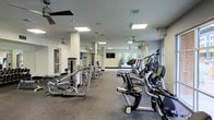 This fitness center offers a comprehensive workout environment designed to accommodate a variety of exercise needs. The space is generously sized, with an open floor plan that allows for easy movement between equipment zones. The flooring is finished with durable rubber matting, ideal for supporting heavy gym equipment and providing traction during workouts.

The cardio section is well-stocked with treadmills, elliptical trainers, and additional aerobic machines, all positioned to take advantage of natural light streaming in through windows along the perimeter walls. A wall-mounted flat-screen television is conveniently placed for entertainment during cardio sessions.

The strength training area features a variety of cable and resistance machines, offering users options for targeting multiple muscle groups. The equipment is arranged to maximize floor space while maintaining clear pathways throughout.

Overhead, recessed flat-panel lighting fixtures provide bright, even illumination throughout the entire facility. Ceiling fans are distributed across the space to promote airflow and comfort during intense workouts. The white walls and light-colored columns contribute to an open, clean aesthetic. A clearly marked exit door ensures safety and accessibility. Overall, the facility presents a practical, well-maintained environment suited for a full range of fitness activities.