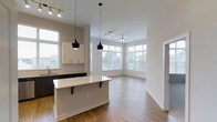 Located at 616 Oberlin Rd in Raleigh, NC, this single-floor space offers a thoughtfully designed open-concept layout that seamlessly connects the kitchen and living areas. The kitchen is a standout feature, showcasing dark cabinetry contrasted by crisp white upper cabinets and a generous island with a light granite countertop and bar seating — ideal for casual dining or entertaining. Pendant lighting in matte black adds a modern, curated touch above the island, complemented by track lighting that illuminates the space with targeted warmth. Hardwood-style flooring extends throughout, providing a cohesive and polished aesthetic. The living area is bathed in natural light, courtesy of expansive floor-to-ceiling windows that wrap around the corner of the room, offering views of the surrounding greenery and cityscape. The high ceilings amplify the sense of openness, giving the space an airy and inviting atmosphere. Neutral wall tones throughout create a versatile backdrop, allowing the architectural details and finishes to take center stage. Overall, this space strikes a balance between contemporary design and functional living, making it a bright and welcoming environment.