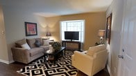This single-floor residence offers a welcoming and well-appointed interior designed for comfortable everyday living. The living room serves as the heart of the space, showcasing a charming bay window that invites natural light to fill the room and creates a bright, open atmosphere. Rich hardwood flooring anchors the space, complemented by a bold black-and-white geometric area rug that adds a striking visual element. Seating is generous and thoughtfully arranged, with a plush gray sofa adorned with decorative pillows and a white armchair providing ample comfort for relaxing or entertaining. A dark wood media console centers the room, housing a flat-screen television framed by the bay window backdrop. Ambient lighting from coordinating table lamps and wall sconces casts a warm glow across the neutral beige walls, enhancing the cozy atmosphere. Decorative touches, including a framed piece of wall art and a lush floral centerpiece on the coffee table, lend personality and polish to the overall aesthetic. The space strikes a balanced blend of modern style and homey comfort, making it an ideal environment for both relaxation and socializing.
