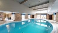 This single-level indoor recreation facility centers around a generously sized swimming pool with vivid blue water, set beneath soaring vaulted ceilings supported by exposed structural beams. Large windows line the perimeter, flooding the pool area with natural light and offering views of the surrounding exterior. The exposed brick walls lend a warm, textured character to the space, contrasting with the crisp white paneling along the upper walls and ceiling.

Adjacent to the pool, a lounge area provides white reclining chaise chairs arranged for relaxation, making it a comfortable space to unwind before or after swimming. The poolside deck features a smooth, easy-to-maintain floor surface. Pendant lighting and recessed fixtures supplement natural daylight, ensuring the space remains well-lit at all hours.

A separate adjoining area, visible through open entryways framed by brick columns, contains dining tables and chairs, suggesting a social or community gathering space. Glass-paneled partitions are also present, offering partial enclosure for additional areas within the facility. Overall, the space combines functionality with a welcoming, leisure-oriented ambiance suited for both recreation and community use.