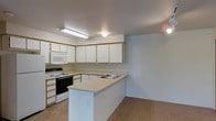 This single-floor space offers a practical and well-organized layout designed for comfortable everyday living. The kitchen serves as a central feature, showcasing a peninsula-style counter that provides additional workspace and separates the kitchen from the adjoining area. White cabinetry lines the walls in an L-shaped configuration, offering generous storage space for both upper and lower cabinet needs. The countertops are finished in a warm neutral tone, complementing the light, clean aesthetic of the space. Essential appliances are included, among them a white electric range with a black oven door, a built-in microwave above the stove, and a double-basin sink integrated into the peninsula. Tile flooring in a warm beige tone runs throughout, adding durability and ease of maintenance underfoot. Overhead lighting, including a fluorescent fixture above the main work area and recessed ceiling lights, ensures the kitchen is well-lit for daily tasks. The walls are painted in a simple off-white tone, creating a bright and neutral backdrop throughout. The open connection between the kitchen and the adjacent space enhances the overall sense of flow and accessibility within this single-level layout.