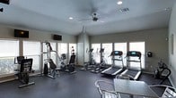 This fitness center is designed to provide a comprehensive workout environment within a single-floor layout. The space is lined with multiple windows fitted with blinds, allowing generous natural light to fill the room while maintaining privacy. A ceiling fan positioned at the center of the vaulted ceiling promotes air circulation, complemented by recessed lighting for consistent illumination throughout.

The cardio section is well-appointed with multiple treadmills featuring digital displays and at least two elliptical cross-trainers, catering to a variety of aerobic exercise preferences. A multi-station cable strength machine and a recumbent exercise bike round out the equipment offerings, supporting both resistance training and low-impact cardio workouts. Medicine balls are also available for functional fitness exercises.

The flooring consists of dark, durable rubber matting — practical for high-traffic fitness use and easy maintenance. A wall-mounted flat-screen television is positioned for visibility during workouts. A stainless steel table and seating near the entrance provide a functional area for rest or personal belongings. The overall ambiance is clean, functional, and well-maintained, making it a practical and inviting space for daily exercise routines.