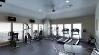 This fitness center is designed to provide a comprehensive workout environment within a single-floor layout. The space is lined with multiple windows fitted with blinds, allowing generous natural light to fill the room while maintaining privacy. A ceiling fan positioned at the center of the vaulted ceiling promotes air circulation, complemented by recessed lighting for consistent illumination throughout.

The cardio section is well-appointed with multiple treadmills featuring digital displays and at least two elliptical cross-trainers, catering to a variety of aerobic exercise preferences. A multi-station cable strength machine and a recumbent exercise bike round out the equipment offerings, supporting both resistance training and low-impact cardio workouts. Medicine balls are also available for functional fitness exercises.

The flooring consists of dark, durable rubber matting — practical for high-traffic fitness use and easy maintenance. A wall-mounted flat-screen television is positioned for visibility during workouts. A stainless steel table and seating near the entrance provide a functional area for rest or personal belongings. The overall ambiance is clean, functional, and well-maintained, making it a practical and inviting space for daily exercise routines.