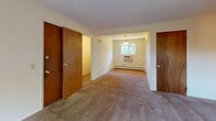 This single-floor unit at 310 N Glen Dr in Rochester, NY, offers a straightforward and functional layout across several well-defined rooms. The floor plan, as seen in the overhead scan, reveals a spacious living area, a separate dining nook, a kitchen, and multiple additional rooms connected by a central hallway lined with warm wood-toned doors.

The living and bedroom areas feature neutral beige walls and wall-to-wall carpeting in a light taupe tone, creating a consistent, understated aesthetic throughout. Baseboard heating units run along the exterior walls in multiple rooms, providing efficient climate control. Windows are positioned throughout the unit to allow ample natural light, with outdoor greenery visible from several vantage points, contributing to a bright and airy feel.

The kitchen is outfitted with dark wood cabinetry, white laminate countertops, light ceramic tile flooring, a black electric range with coil burners, a white refrigerator, and a stainless steel sink. A wall-mounted ventilation fan is positioned above the range. The kitchen opens into the dining area, which features a pendant light fixture and a window with a wall-unit air conditioner below it, complementing the baseboard heating system found elsewhere in the unit.