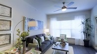 This single-floor residence centers around a thoughtfully designed living area that balances comfort and contemporary style. The living room is highlighted by soaring ceilings that lend an open, airy feel, complemented by a ceiling fan with integrated lighting that enhances both airflow and illumination. A large window dressed with sheer white curtains and horizontal blinds allows generous natural light to fill the space, creating a warm and inviting atmosphere throughout the day. The walls are finished in a soft, neutral tone that serves as a versatile backdrop for décor. Built-in or wall-mounted art adds a splash of color, while the carefully selected furnishings — including a plush dark sofa adorned with green and white accent pillows and a coordinating accent chair — contribute to a cohesive design aesthetic. A wood-finished coffee table anchors the seating area over a textured area rug, adding warmth underfoot. The space is accented with greenery and natural decorative elements, enhancing its fresh, modern appeal. Dark hardwood-style flooring runs throughout, providing an elegant contrast to the light walls and furnishings.