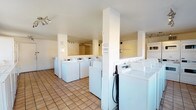 This shared laundry facility is a practical, well-organized space designed to accommodate multiple users simultaneously. The room is lined with a generous number of top-loading washing machines and dryers, arranged in rows and sections divided by white structural columns that add a clean, orderly look to the layout. The warm-toned ceramic tile flooring is durable and easy to maintain, running throughout the entire space. The walls are finished in a soft cream hue, contributing to a bright and welcoming atmosphere. Overhead, track lighting provides ample illumination across the room, ensuring good visibility at all times of day. A central open area between the machine rows allows for comfortable movement and easy access to all units. Posted signage on the walls provides usage instructions and facility guidelines for users. A white entry door is visible at one end of the room, offering straightforward access to the facility. The space is thoughtfully laid out to maximize the number of machines while maintaining clear circulation paths, making it a highly functional communal laundry room suited for a multi-unit residential building or similar shared-use property.
