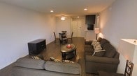 This single-floor space is designed with an open-concept layout that seamlessly integrates the living and dining areas into one cohesive environment. The living area is anchored by plush, dark upholstered sofas arranged around a round coffee table with a decorative tray centerpiece, creating an inviting seating space. A wall-mounted flat-screen television is positioned on an elevated shelf, complemented by a media console along the adjacent wall, making the area both functional and entertainment-ready. A floor lamp provides warm accent lighting alongside recessed ceiling lights that illuminate the space evenly throughout.

The dining area, situated just beyond the living space, features a round table paired with dark wooden chairs, offering an intimate setting for meals. Hardwood-style flooring runs throughout, providing a warm and cohesive aesthetic that ties the areas together. A ceiling fan with a light fixture serves as a practical central feature, enhancing airflow and comfort. Neutral-toned walls and clean architectural lines contribute to the space's understated, modern ambiance. A doorway at the far end suggests additional rooms or access points beyond the main open living area.
